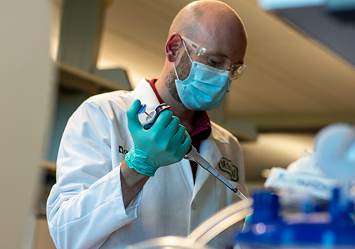 Person in lab wear, safety goggles, and mask working with pipette. 
